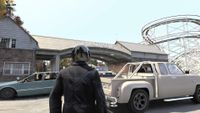 A GTA IV character in a dark jacket and motorcycle helmet standing in front of cars, with a roller coaster in the background.
