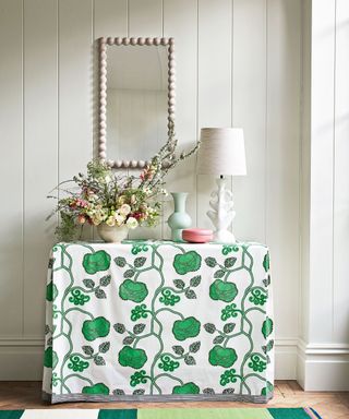 console table in hallway with green and white floral table cloth, bobbin framed mirror on white wall, cream table lamp, vase of flowers