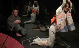 View of a performance featuring Japanese rope bondage - there is a red parasol in the corner, a man in a dark patterned robe sitting on the floor and a woman in a light coloured floral outfit and orange waist tie kneeling with her hands bound by rope above her head