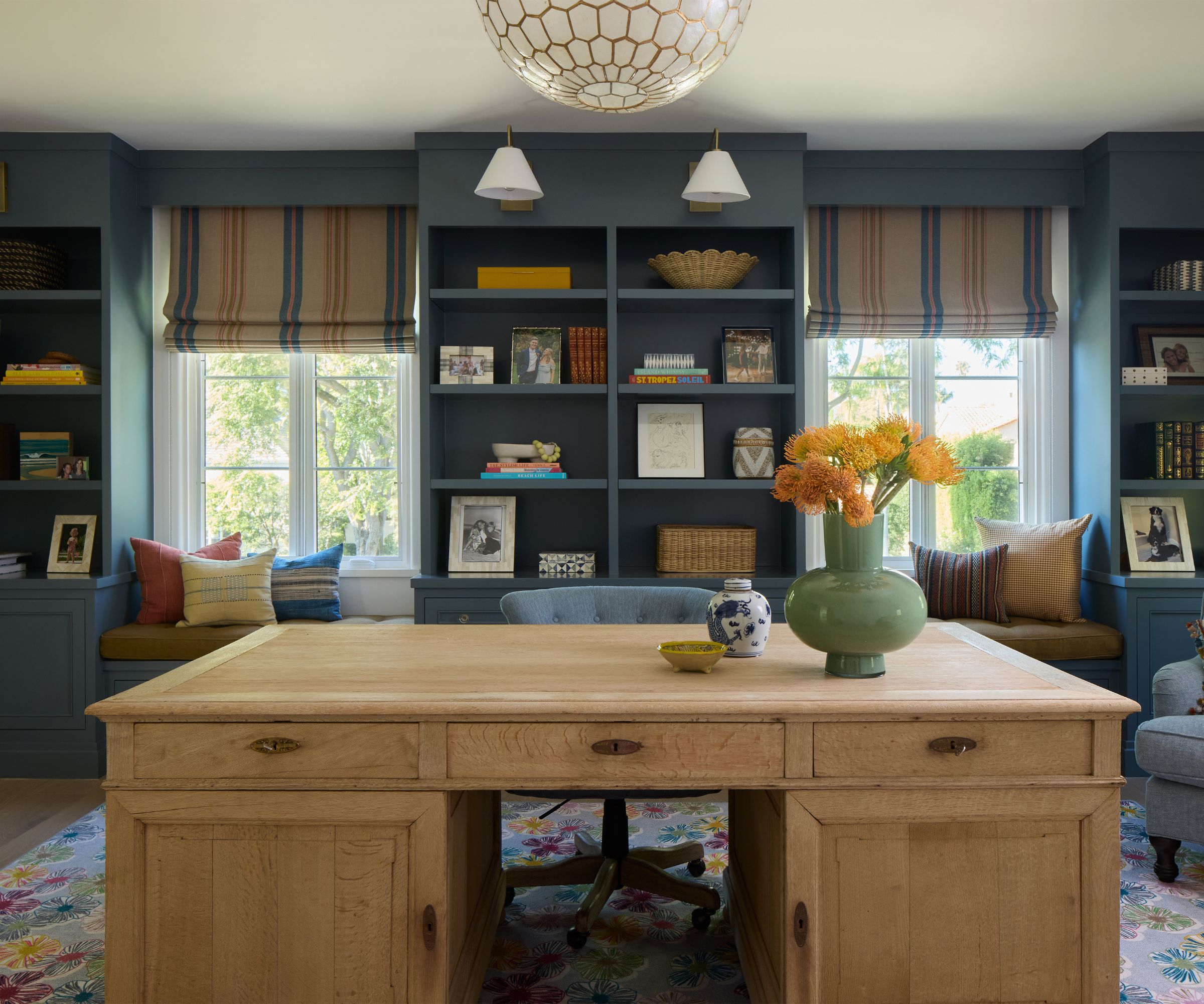 Home office with blue painted walls and built-in shelving, window seats, wood desk and green vase with yellow flowers