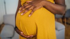 Close-up photo of a Black woman who is wearing a yellow dress and is holding her hands around her pregnant belly. 