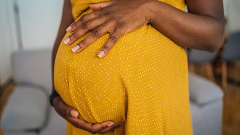 Close-up photo of a Black woman who is wearing a yellow dress and is holding her hands around her pregnant belly. 