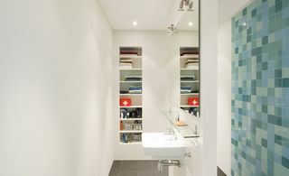 Bathroom with white walls. grey flooring and a featured wall with squares in shades of blue. An inbuilt shelf with 8 levels. White handwash sink with a mirror above
