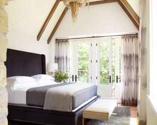 monochrome bedroom with vaulted ceilng with beams and shiplap, black bed with winged headboard, end of bed bench, patterned curtains