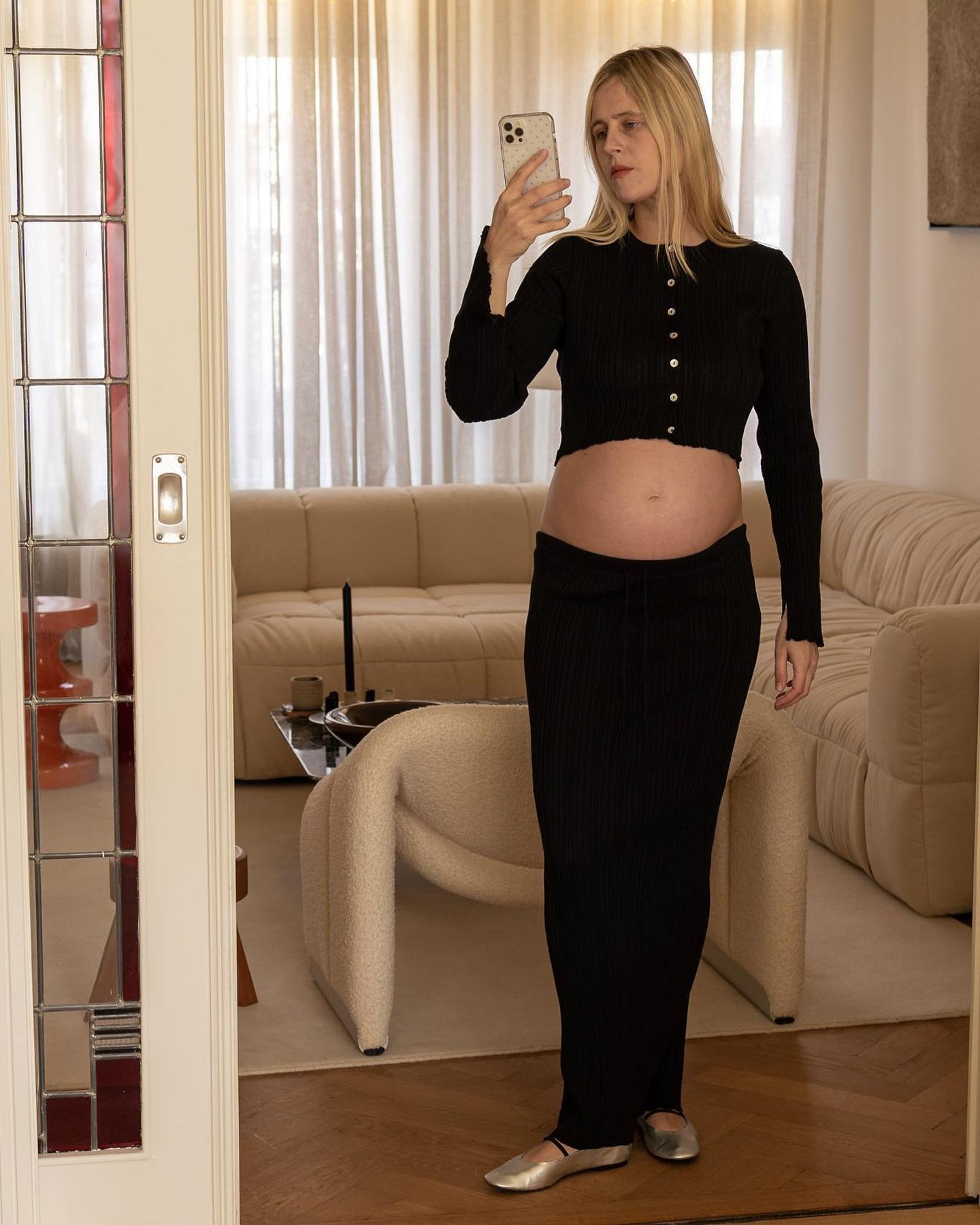 Dutch style influencer Linda Tol poses in her Amsterdam living room wearing a maternity outfit with a black cropped button-front cardigan, a black ribbed maxi skirt, and sliver Mary Jane flats
