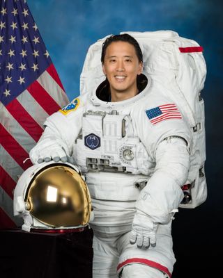 jonny kim in a spacesuit with a helmet by his side and the american flag in behind