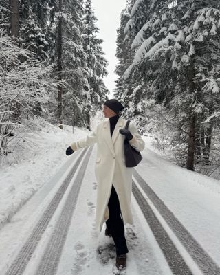 Mary is wearing a black beanie hat, long white coat, black turtleneck sweater, black leather tote, black pants, and black boots.