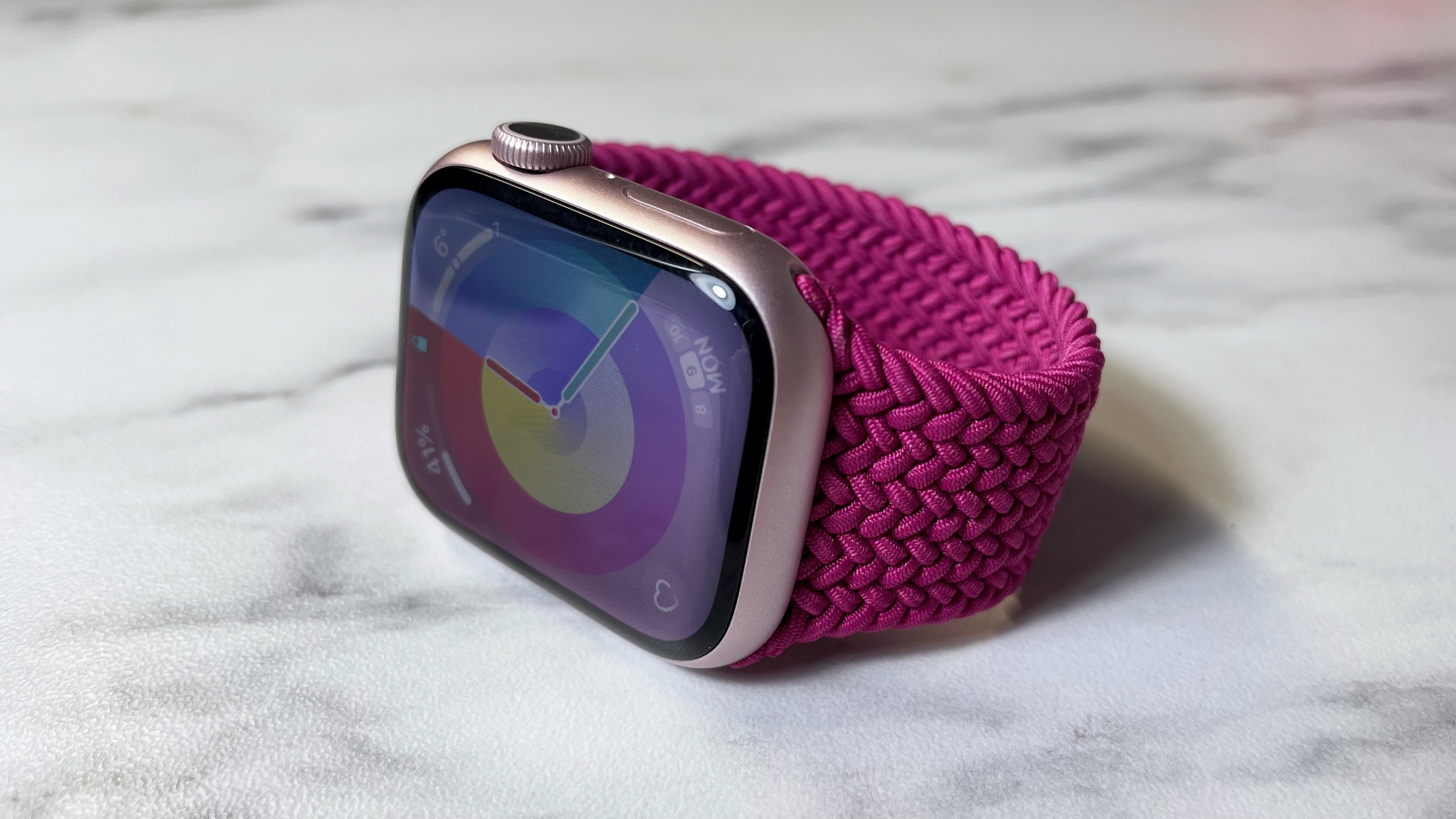 A magenta-colored Apple Braided Solo Loop is attached to a light pink Apple Watch angled slightly to the left on a marble-effect surface.