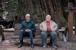 Two men (Ted Danson as Charles, Stephen McKinley Henderson as Calbert) sit on a wooden bench in front of a giant true trunk, with a tall cross to their right, in episode 107 of 'A Man on the Inside.'