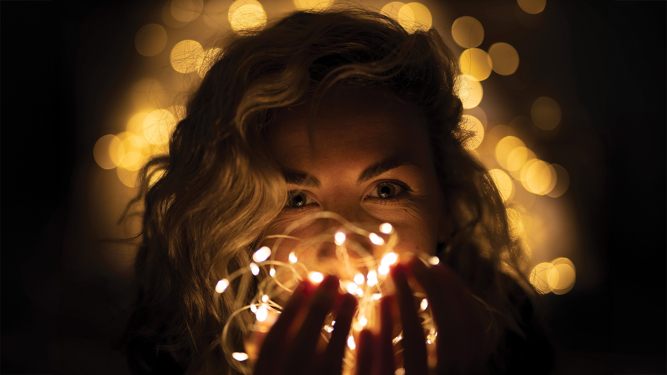 A woman with curly hair holds glowing fairy lights close to her face, creating a warm, magical ambiance. Her eyes sparkle with joy against a blurred background of soft bokeh lights