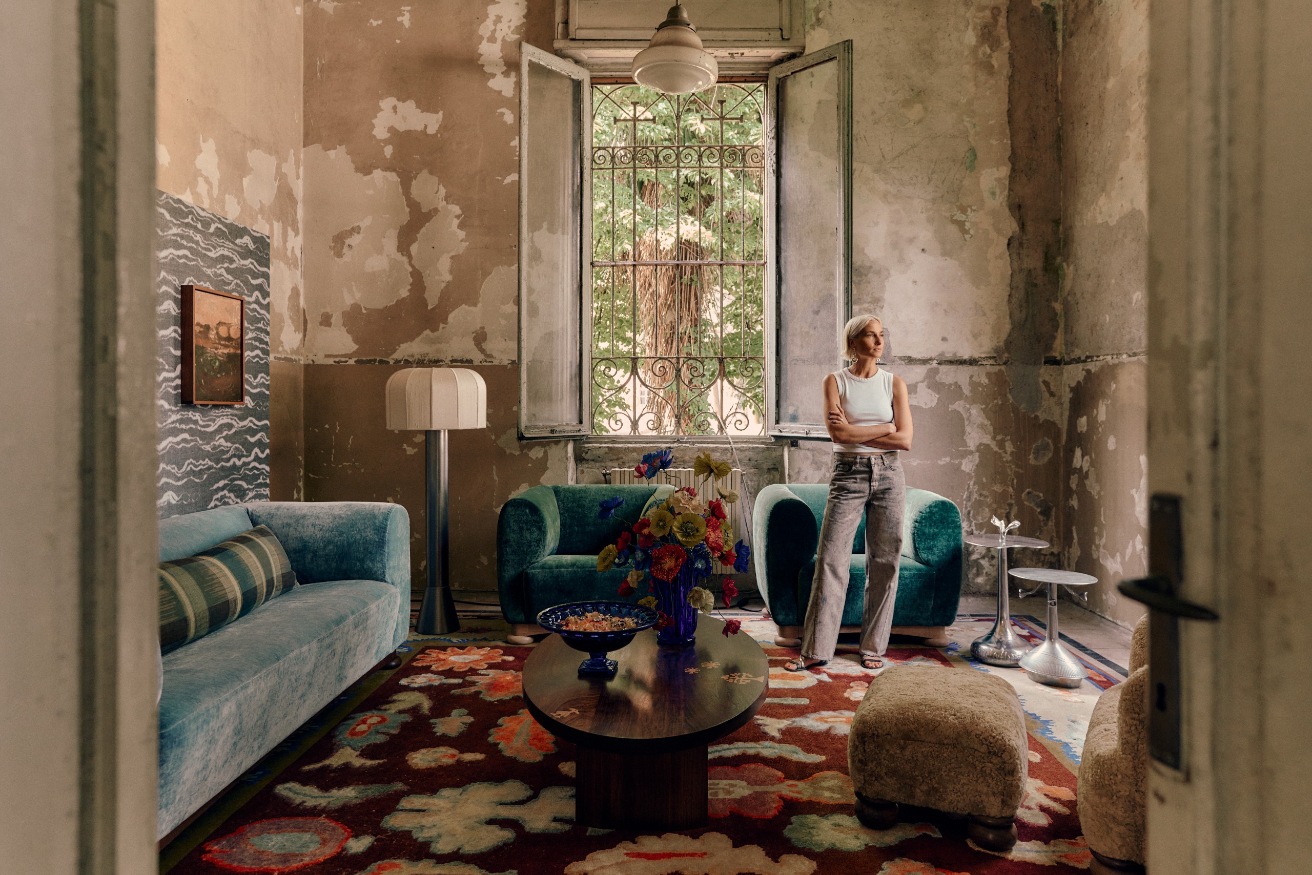 A young woman in a white tank top with gray trousers stands in a layeredly decorated room withbare cement walls and plush, velvety furniture with her arms crossed.