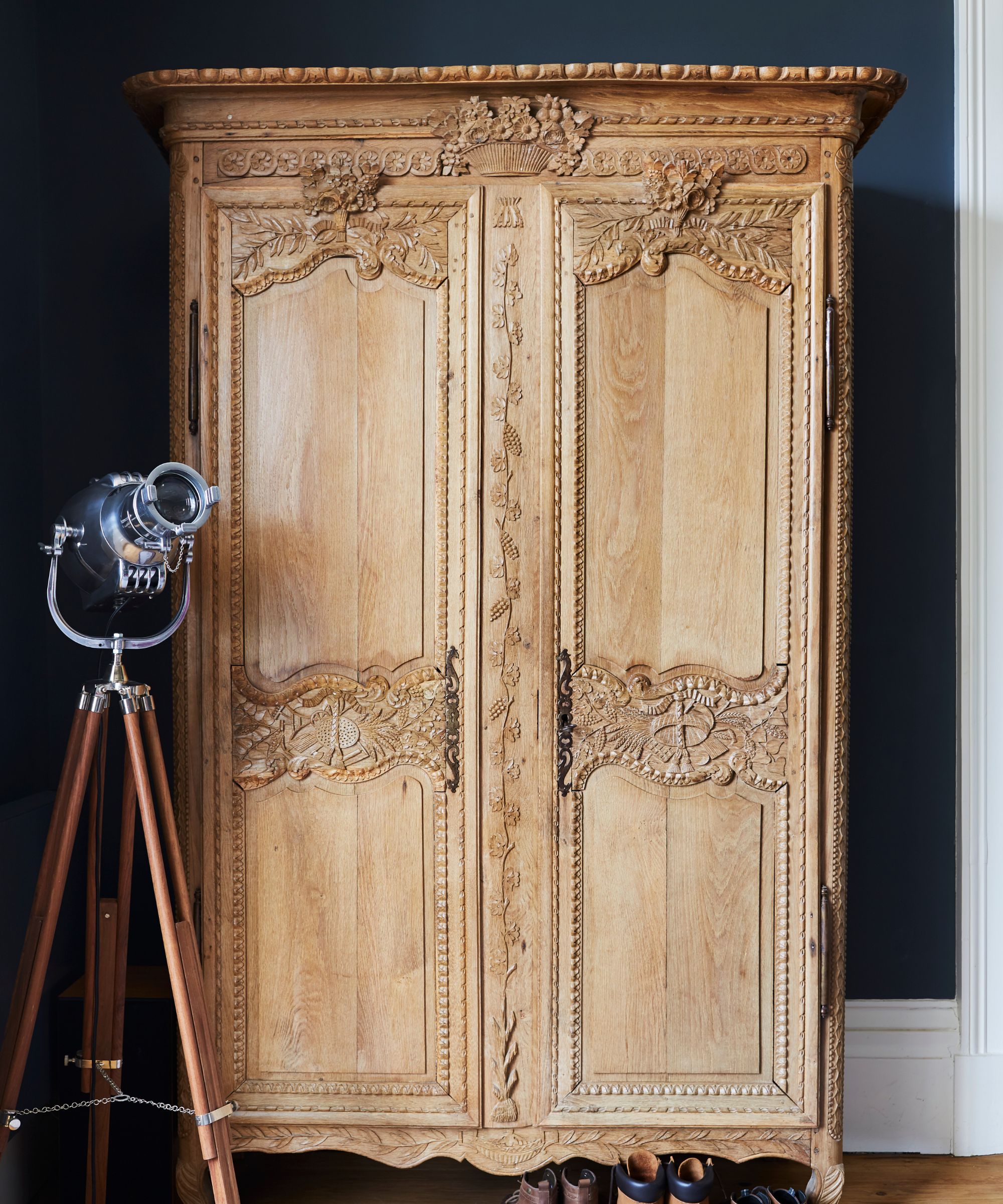 A wooden carved wardrobe with dark walls behind, white baseboards, a free-standing statement lamp and wooden flooring.