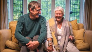 Louis Theroux and Judi Dench laughing sat together on a sofa.