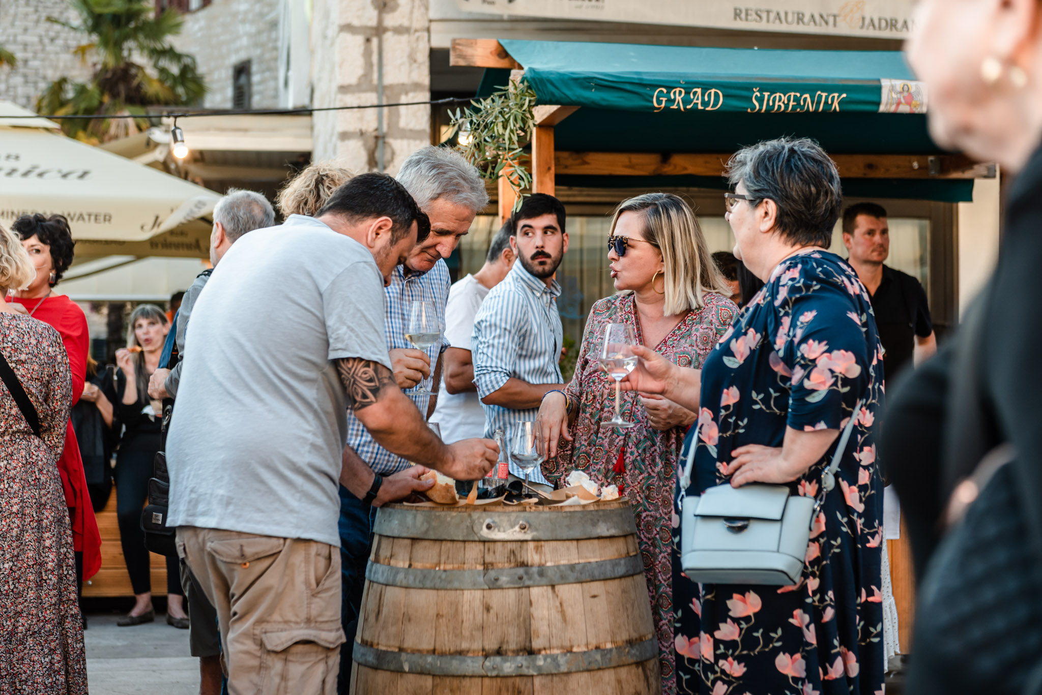 People standing around a wine barrel, holding glasses of white wine and eating food