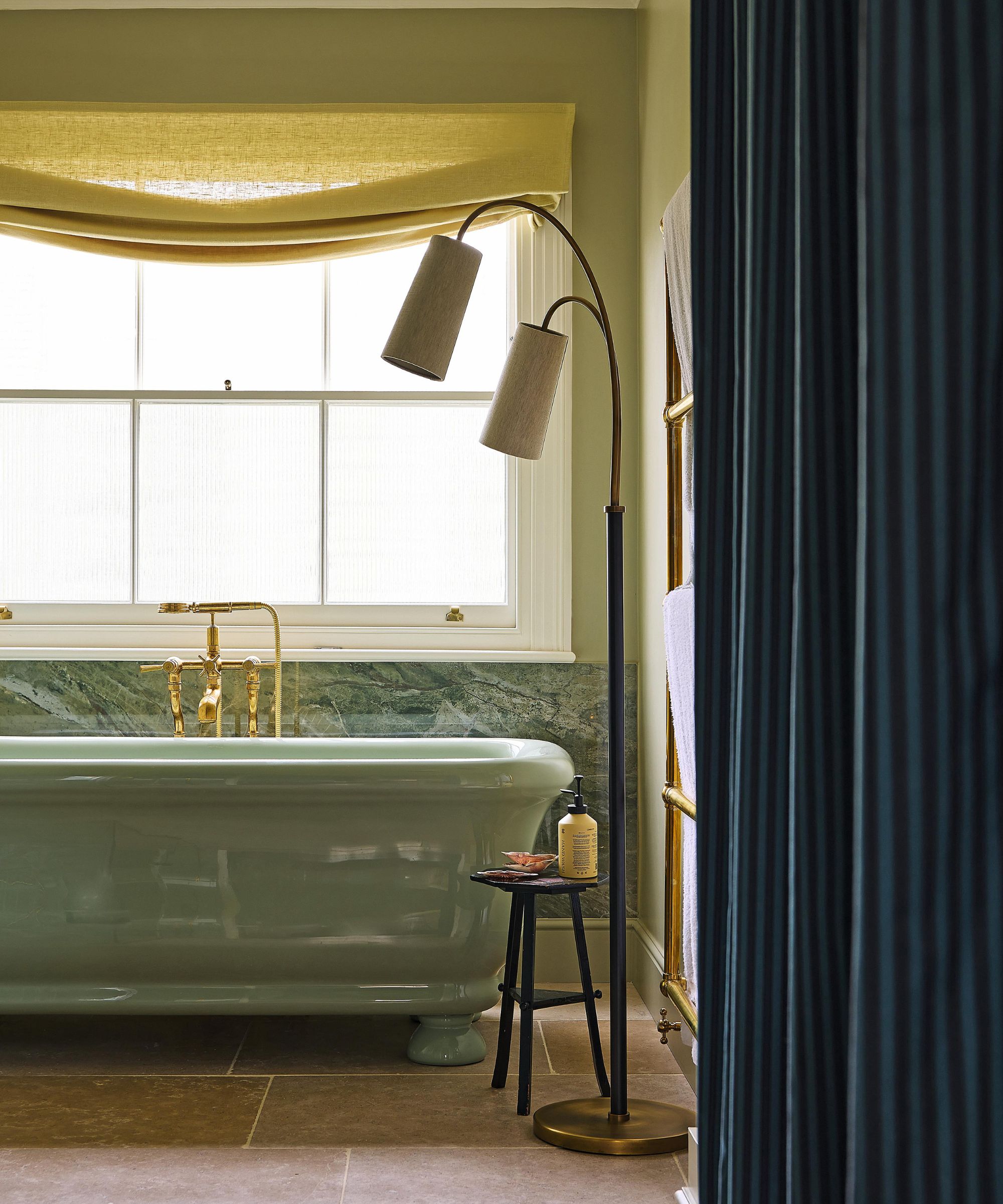 a london bathroom with soft green walls, a yellow relaxed roman blind, green marble tile halfway up the wall, and a freestanding green bathtub