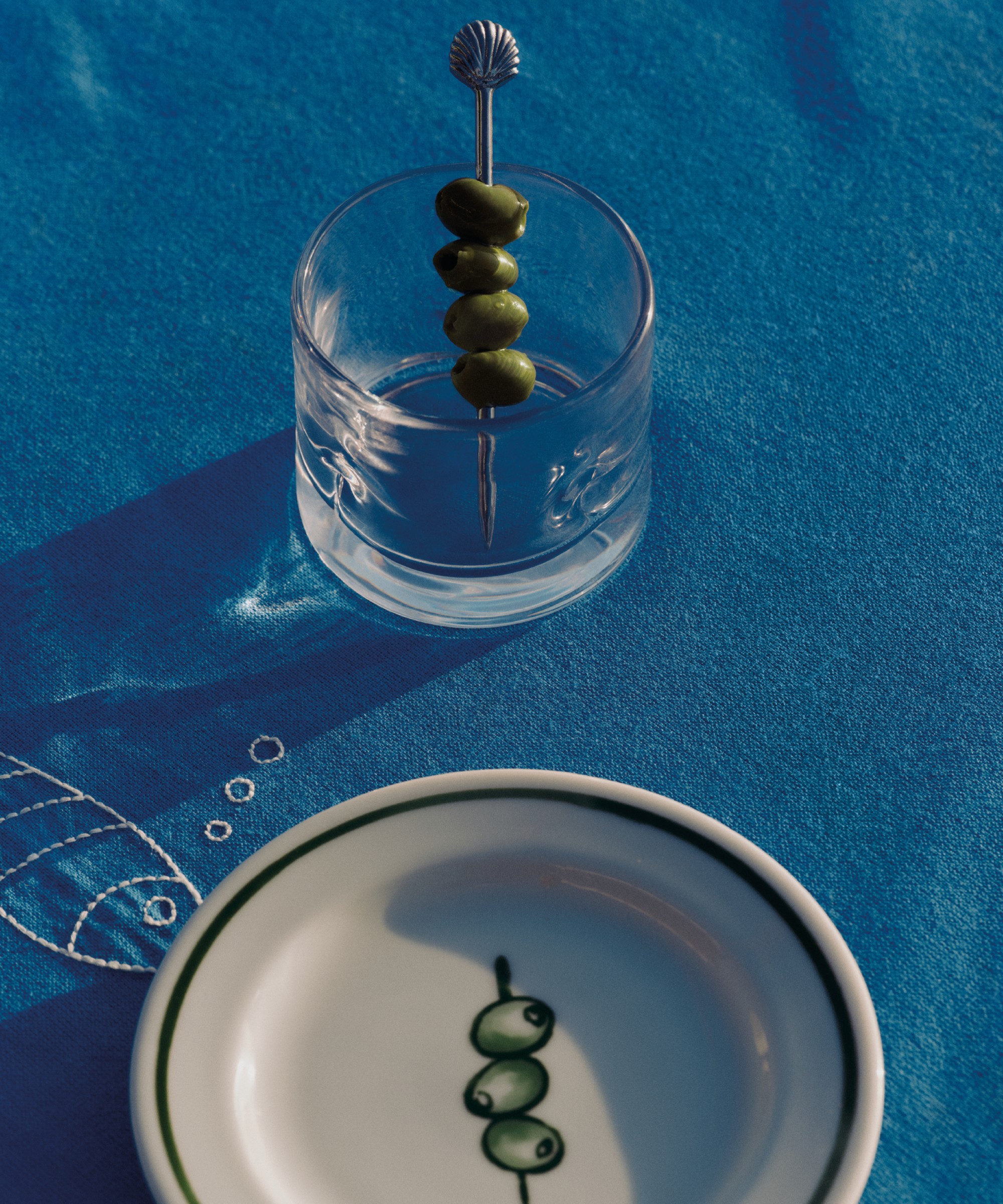 A tumbler glass from H&amp;amp;M Home's Riviera-themed summer 2026 collection with a shell cocktail pick with olives and a side plate with an olive print