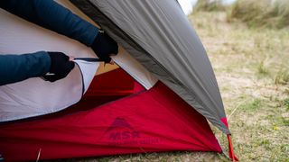 The corner of a MSR Hubba Hubba Bikepack One-Person Tent, pegged out on a grassy, sandy beach. A pair of hands are reaching across and opening the tent door from the outside.