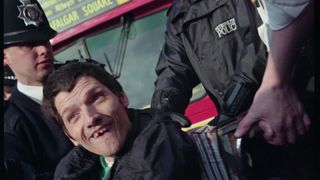 A wheelchair user in a pink sweatshirt holds a sign reading "How about us?" beside a red London Leyland bus, as bystanders look on from the pavement.