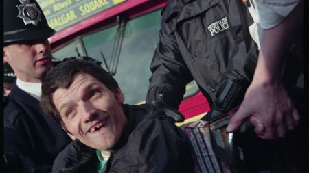 A wheelchair user in a pink sweatshirt holds a sign reading "How about us?" beside a red London Leyland bus, as bystanders look on from the pavement.