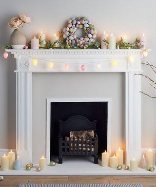 A white fireplace with pastel colored candles and eggs on top of it and on the edges of it, with a mantel with a ceramic vase of flowers, a green floral garland, and an egg wreath and egg string lights