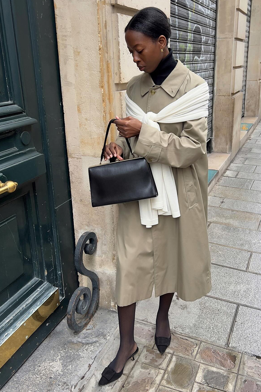 Layering staples for spring 2026 is shown in a street-style photo of a woman standing outside, wearing a black turtleneck sweater under a tan trench coat, with a white scarf tied around the shoulders. The rest of the look is styled with a black skirt, black sheer tights, black mules, and a black frame handbag