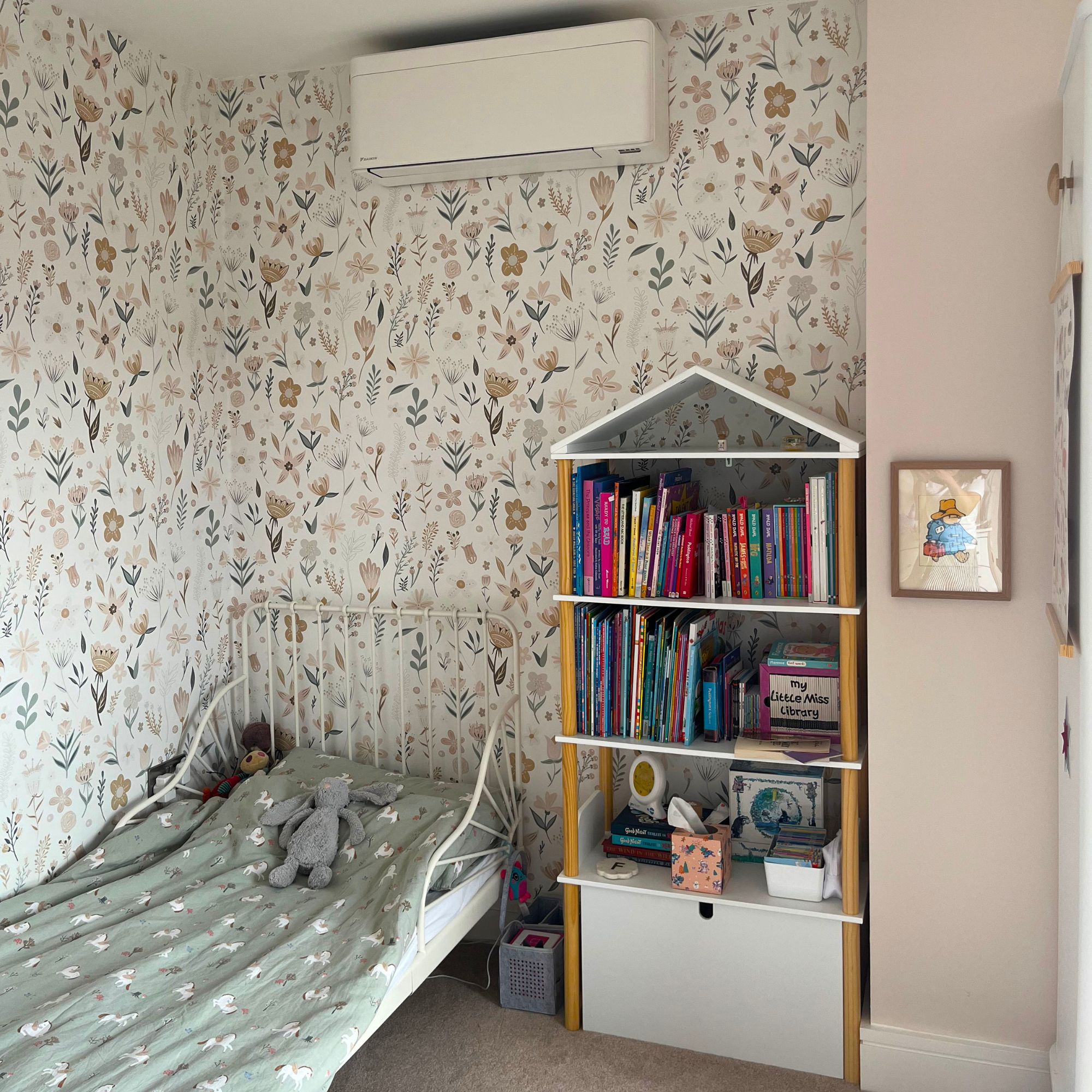 Fitted air conditioning unit on a wallpapered wall in a child's room above a low bed and house-shaped bookcase