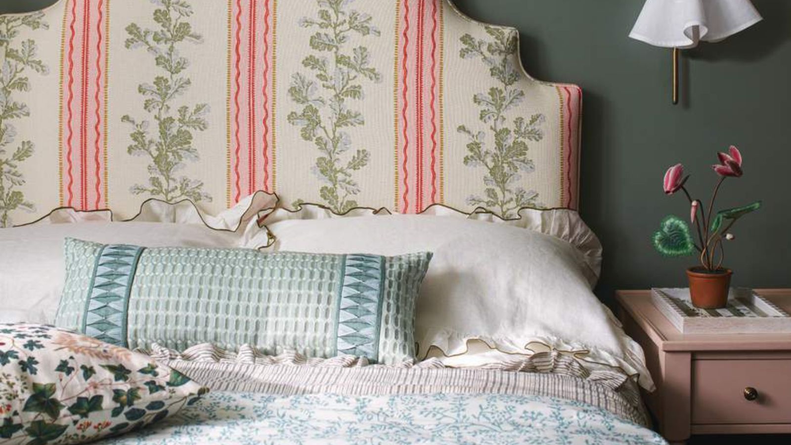 White and blue bedding on a pink, green and white headboard against a green wall; to the right, a vase of flowers on a nightstand.
