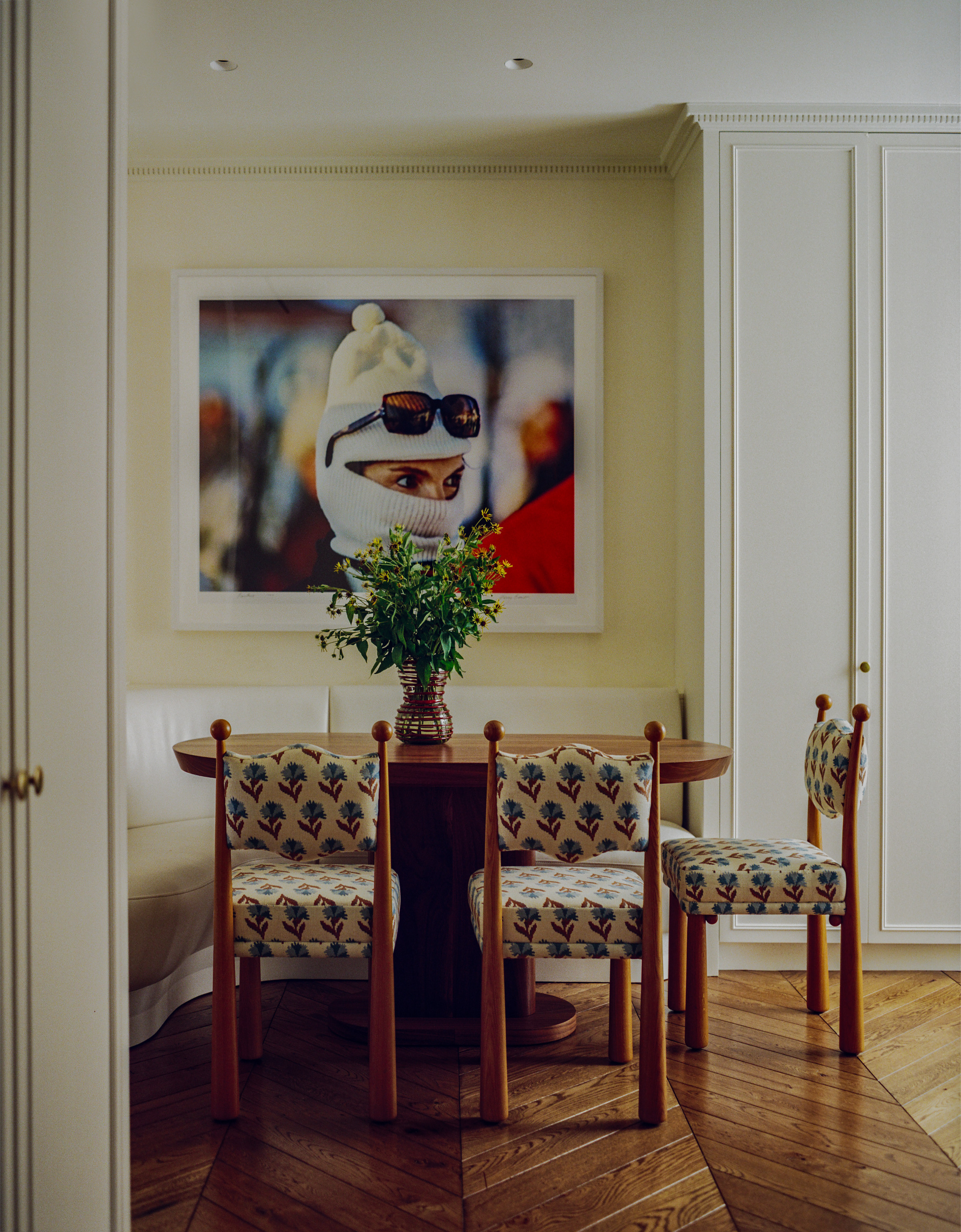 Butter yellow breakfast area with cream curved banquette seating, wood table, wood and floral pattern chair and oversized Jackie Onassis ski picture