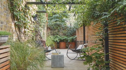 Raised, tiled seating area with black pergola and greenery above, and wooden fencing along the length of the outdoor space. 