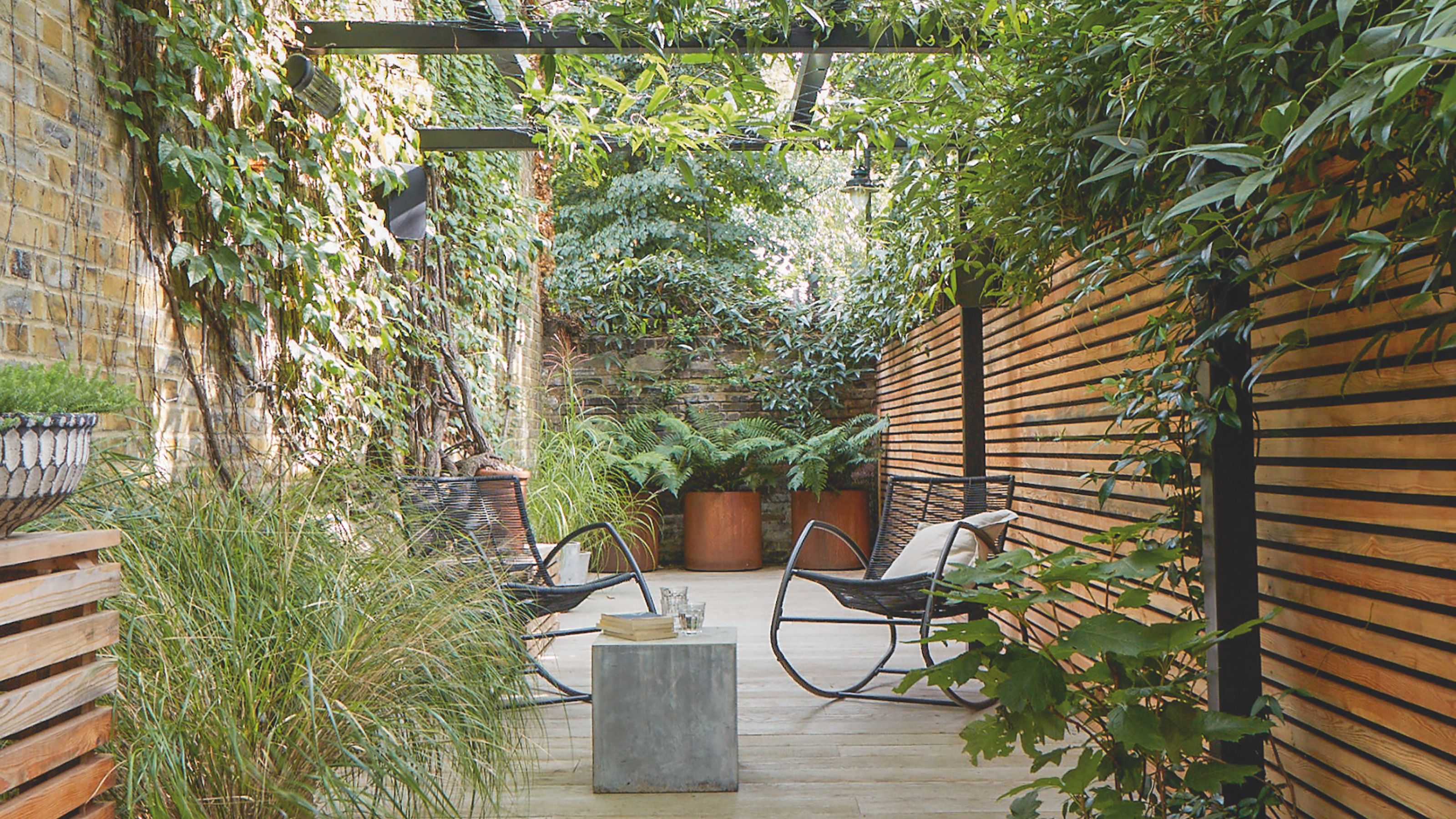 Raised, tiled seating area with black pergola and greenery above, and wooden fencing along the length of the outdoor space. 