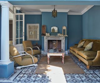 blue and white patterned floor tiles in living room space with blue walls, mustard colour sofas, blue columns and white rustic fireplace