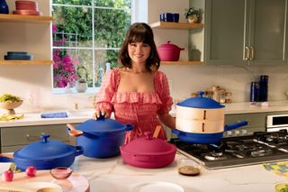 Selena Gomez standing in front of a kitchen island with blue and pink pots and pans balanced on top