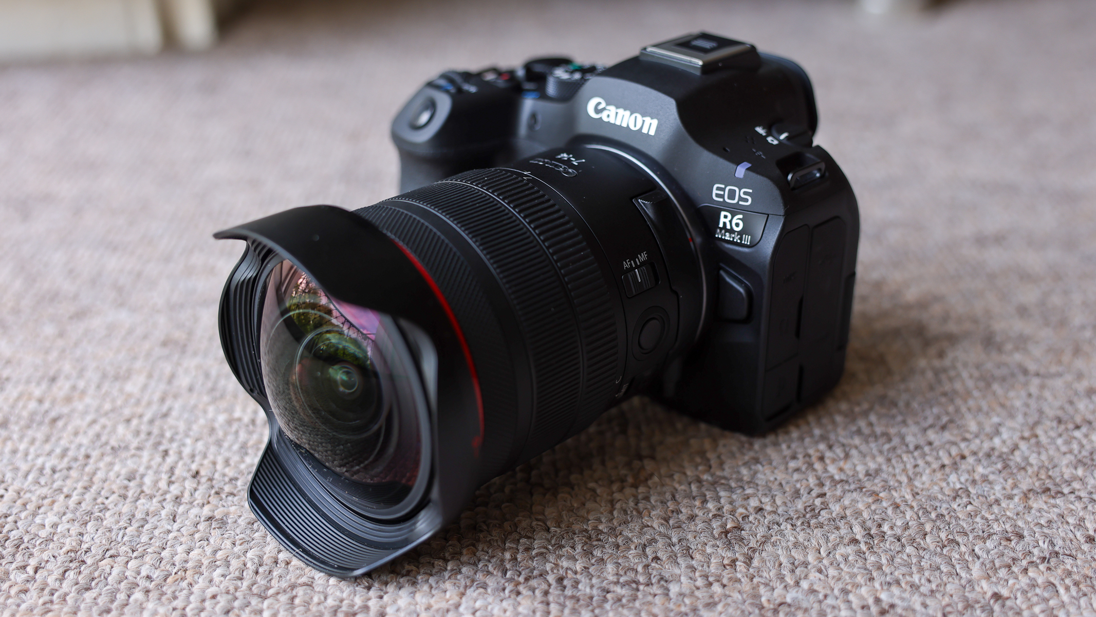 The Canon EOS R6 III with the Canon RF 7-14mm f/2.8-3.5L fisheye lens attached, on a brown carpet.