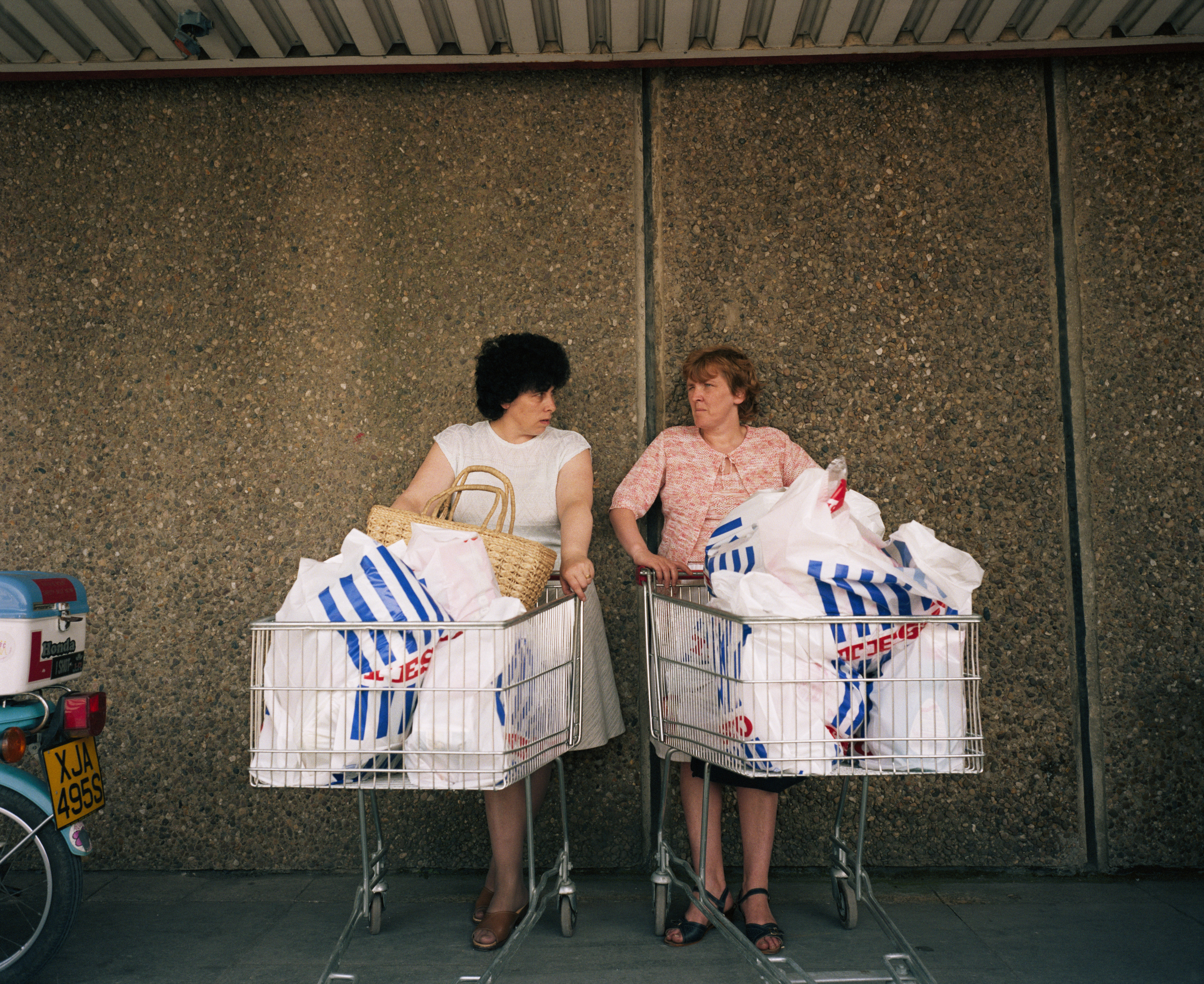 Two women stand side-by-side against a pebbled wall, each leaning on a shopping cart overflowing with white and blue plastic grocery bags.