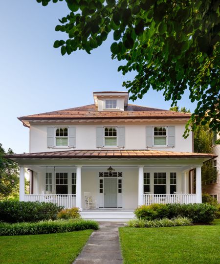 A home exterior with white siding, light blue window shutters and front door, a red brick roof and a full-length porch and grass lawn. 