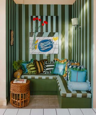 Bold and eclectic corner banquette featuring green and blue striped upholstery, vibrant patterned cushions, striped wallpaper, and colourful artwork for a playful, maximalist look.