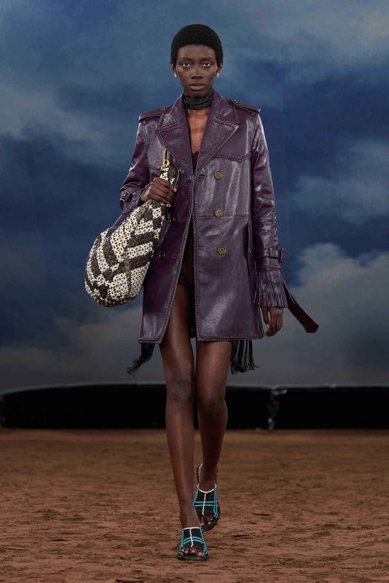 A Burberry model wearing an aubergine-hued leather jacket, a black bodysuit, a black fringe scarf, woven blue and white mules, and a woven brown and white leather bag.