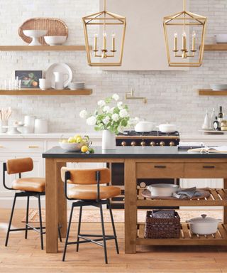 Two terracotta stools around a wooden kitchen island, topped with a vase filled with flowers; in the background, white kitchen cabinets and wooden shelves; two gold pendant lights hanging from the ceiling.