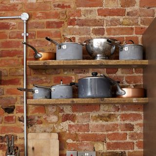 A kitchen with a red brick wall and shelving filled with pots and pans