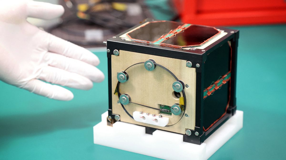 closeup photo of a small cubical brown satellite sitting on a green table, with a white-gloved hand next to it for scale