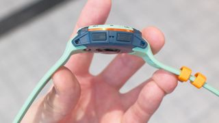 Close-up side view of Garmin Instinct 3 GPS smartwatch in Neotropic/Twilight with a grey and orange case and sea foam green strap. Two buttons are visible on the case