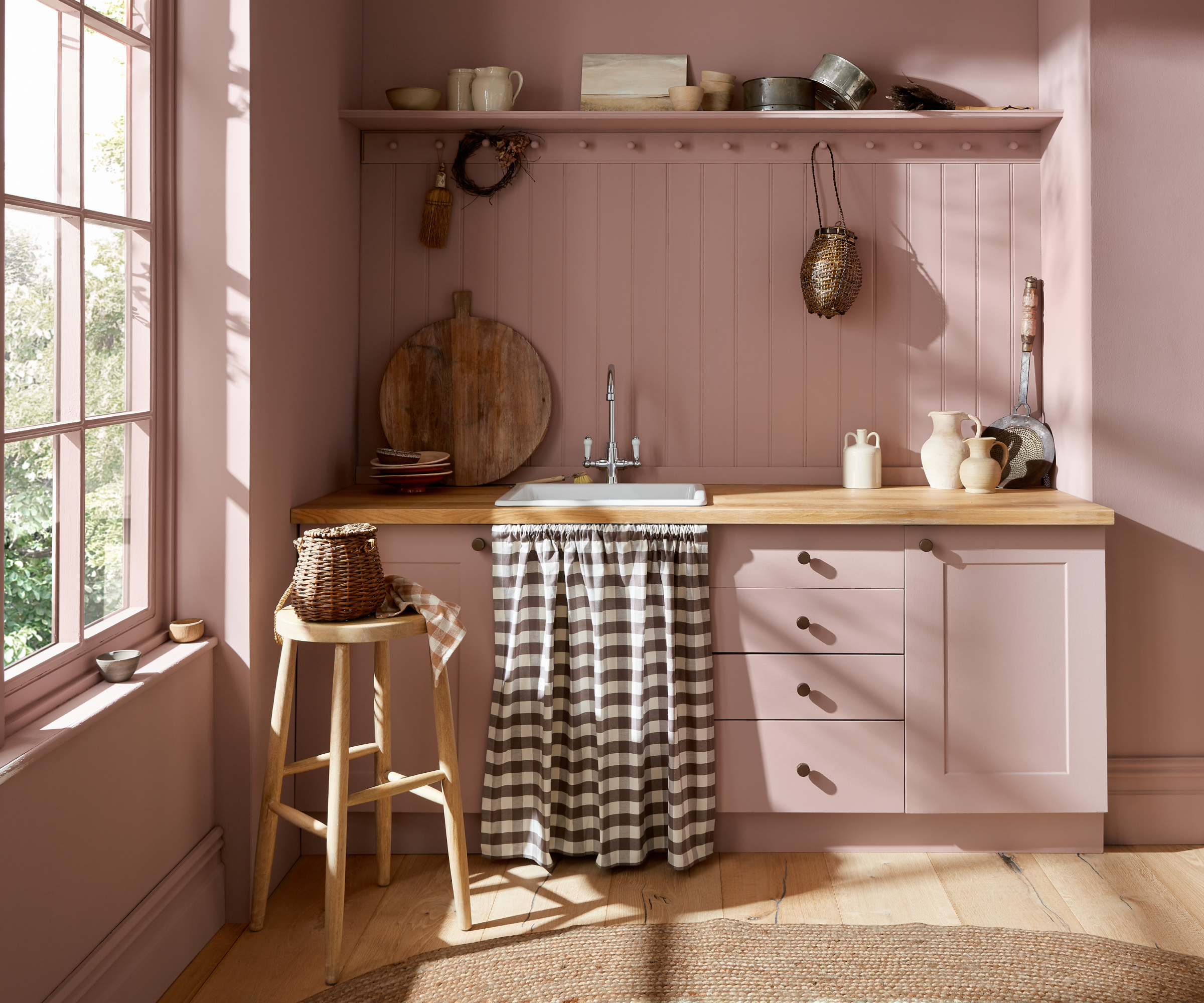 small kitchen with large sash window, wooden flooring, and cabinets, window frame, walls and panelling painted in same shade of pink