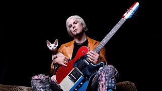 John 5 seated, holding a guitar, with a hairless cat looking over his shoulder