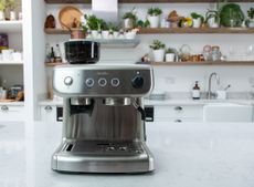 Breville Barista Max espresso machine review by Real Homes. The stainless steel like item is pictured on white marble kitchen counter with open wooden shelving in soft focus in background