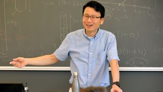 A smiling professor in a button-up blue shirt teaching in front of a chalkboard