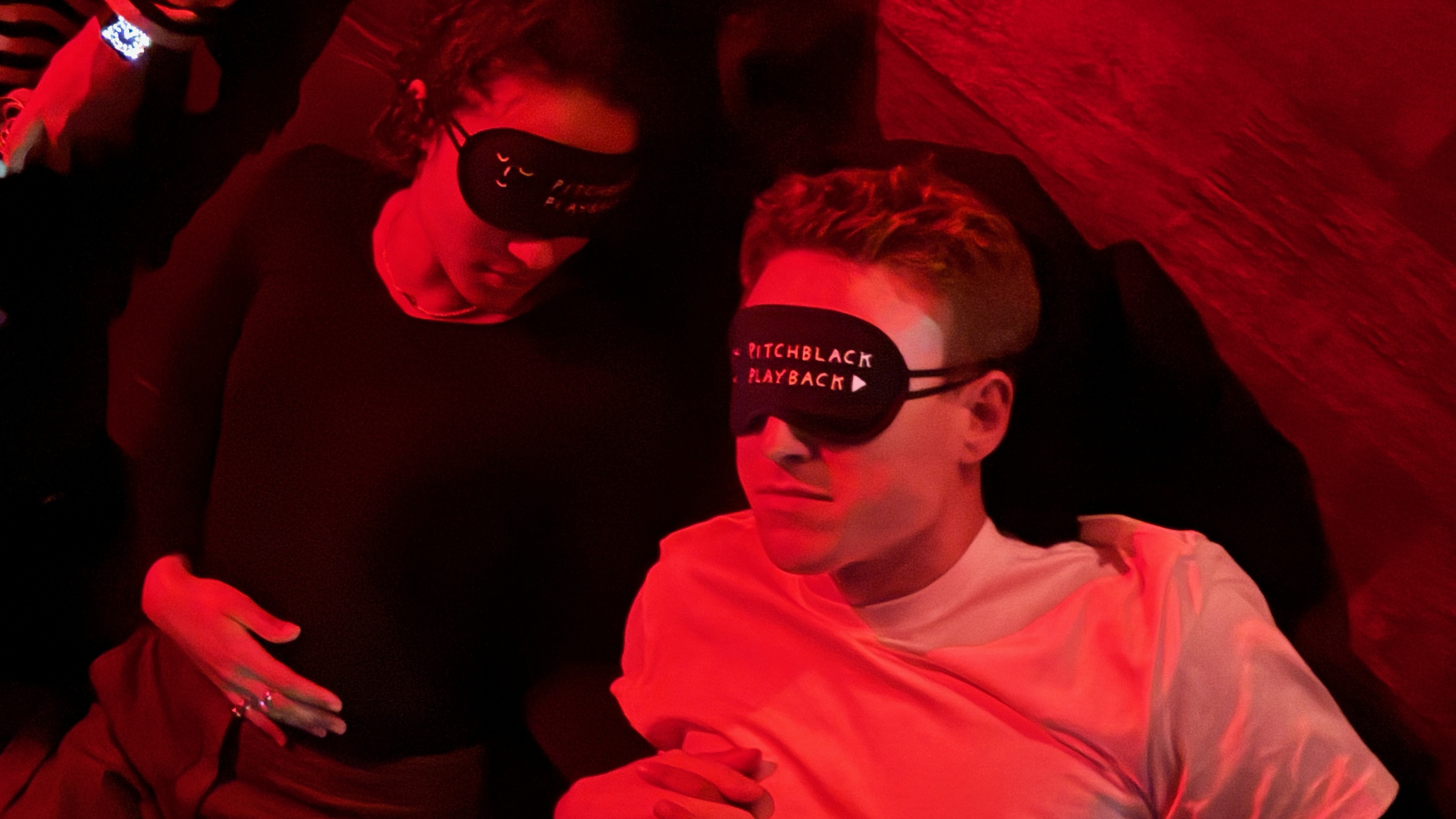 A couple wearing Pitchblack Playback eye masks holding hands in the dark.