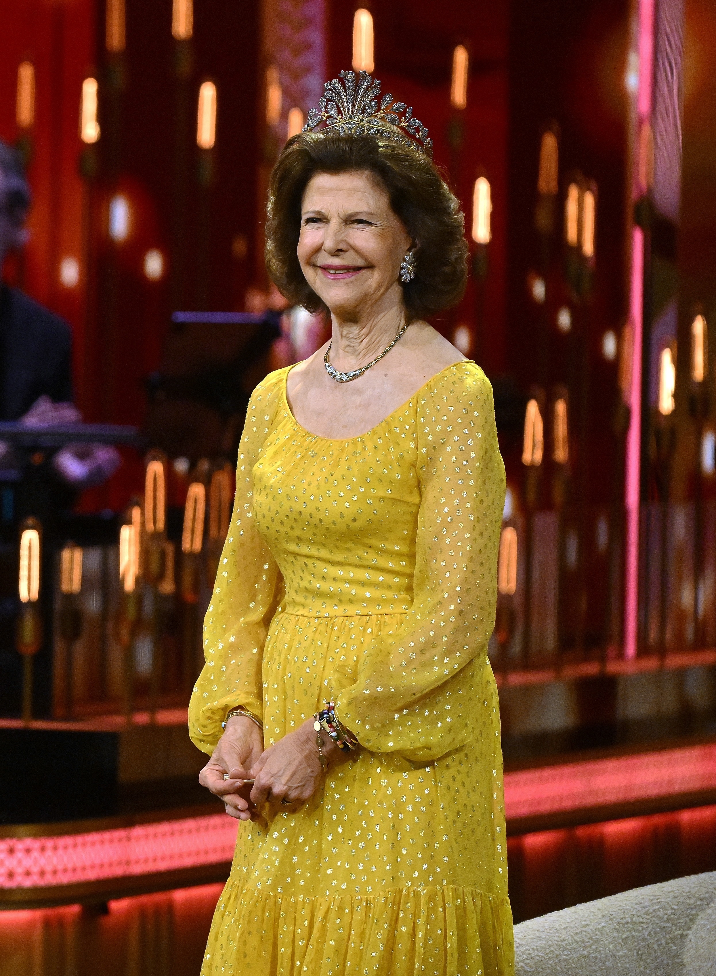 Queen Silvia wearing a yellow Dior gown and tiara on stage