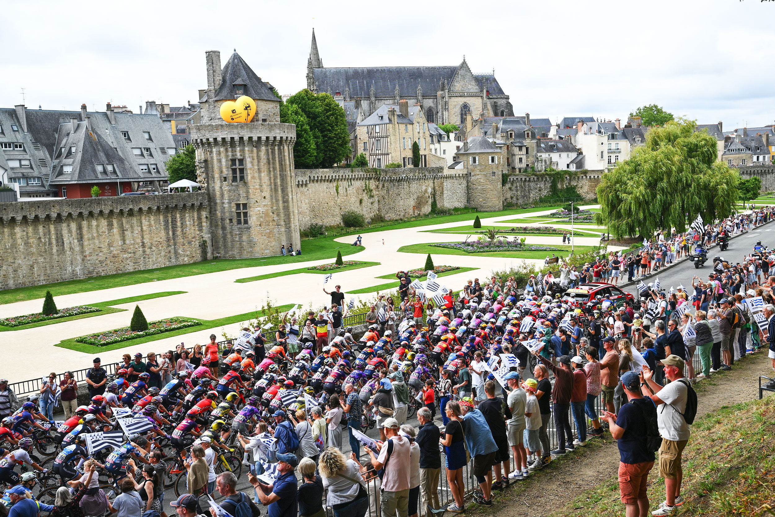 A wide-angle shot of the Tour de France Femmes peloton riding past packed Breton fans and the ancient city walls of Vannes during stage 1 of the race