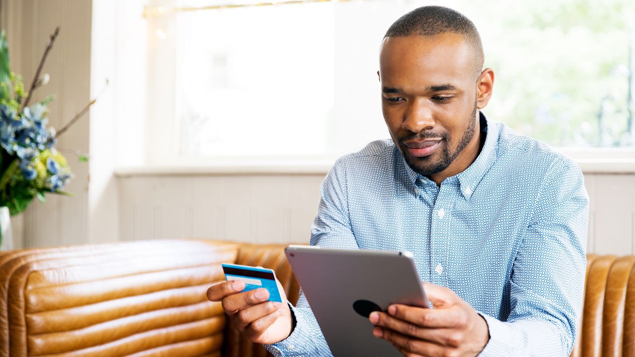 A man holds a credit card in one hand while he looks at a tablet in the other.