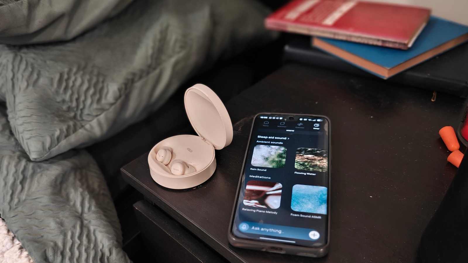 The Fitnexa SomniPods 3 on a bedsite table next to books, earplugs and a smartphone showing the Fitnexa app.